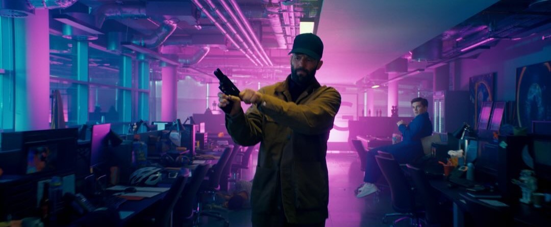 Adam Clay is standing inside of a long room, with computers on each side of him. The lights are bright purple and blue, and they reflect off of him. He is cocking his gun and staring menacingly.