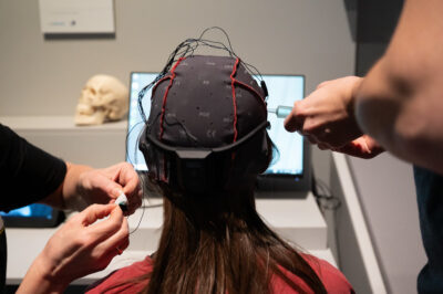 A person looking at a computer screen with a brain scanning cap on and two pairs of hands manipulating the wires on the cap.